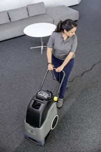 Person using a Kärcher carpet cleaner in an office setting with grey carpet, a sofa, and a round white table in the background.
