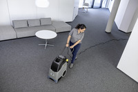 Person using a Kärcher carpet cleaner in a modern office with grey carpet, a grey sofa, and a round white table.