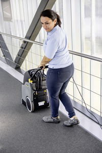 Person using a Kärcher carpet cleaner on a grey carpeted floor in a modern indoor setting with glass walls.