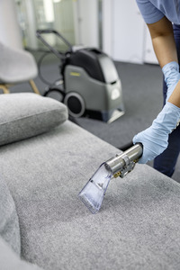 Person cleaning a grey sofa with a Kärcher carpet cleaner in an office setting, wearing blue gloves.