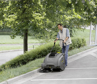 Person operating a Kärcher sweeper on a paved path next to grass and trees in an outdoor setting.