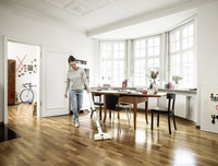 Person cleaning wooden floor with Kärcher electric broom in a bright room with large windows, a table, chairs, and decorative items.