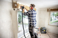 Person sanding wall near window using a power tool, standing on a ladder in a room with unfinished walls. Kärcher vacuum cleaner on the floor.
