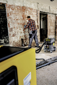 Person in plaid shirt working on brick wall with Kärcher vacuum cleaner in construction setting. Yellow equipment in foreground.