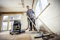 Person cleaning a dusty room with a Kärcher vacuum cleaner, standing near large windows with a view of a tiled roof.