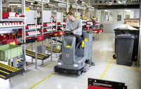 Person operating a Kärcher ride-on floor scrubber in a warehouse with shelves of red bins and industrial equipment.