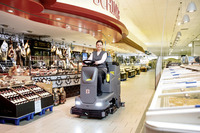 Person operating a Kärcher ride-on floor scrubber in a supermarket aisle, surrounded by deli meats and wine displays.