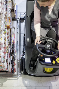 Woman operating a Kärcher ride-on floor scrubber near a supermarket shelf stocked with various packaged goods.