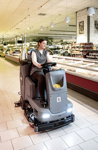 Person operating a Kärcher ride-on floor scrubber in a supermarket aisle, surrounded by shelves of products.