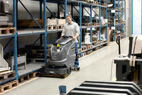 Person operating a Kärcher floor scrubber in a warehouse with metal shelving and industrial equipment.