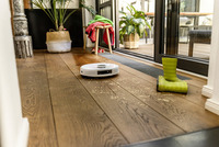 White robotic vacuum on wooden floor in a sunlit room, near green boots and indoor plants by a glass door.