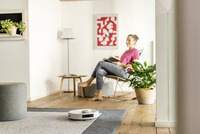 Living room with a robotic vacuum on a rug, a person reading in a chair, potted plant, and abstract red artwork on the wall.