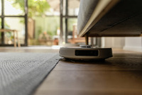 Robotic vacuum cleaner navigating under a sofa in a sunlit living room with wooden floors and a grey rug.