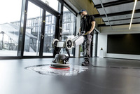 Person operating a Kärcher floor scrubber on a shiny indoor surface, with large glass windows and modern architecture in the background.