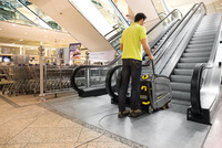 Person operating a Kärcher escalator cleaner in a shopping centre near escalators and shopping trolleys.