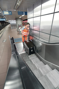 Worker in orange vest operates Kärcher escalator cleaner in a subway station, with signage and tiled walls visible.