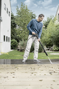 Man cleaning wooden deck with Kärcher high-pressure cleaner in a garden setting, surrounded by greenery and adjacent buildings.