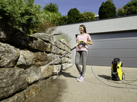 Person using a Kärcher high-pressure cleaner to wash a stone wall outdoors, with a modern garage and greenery in the background.