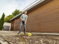 Person cleaning outdoor patio with Kärcher surface cleaner, standing near modern wooden panelled building.