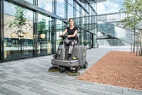 Person operating a Kärcher ride-on sweeper on a paved area outside a modern glass building with trees and gravel landscaping.