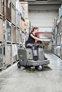 Person operating a Kärcher ride-on floor sweeper in a warehouse aisle, surrounded by stacked metal cages and boxes.