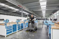 Person operating a Kärcher ride-on sweeper in a spacious industrial workshop with workbenches and blue vices.