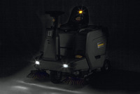 Kärcher ride-on floor sweeper with headlights on, featuring rotating brushes, in a dark setting.