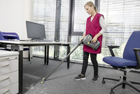 Person vacuuming office carpet with Kärcher cordless vacuum cleaner, near desk and blue office chair, large windows in background.