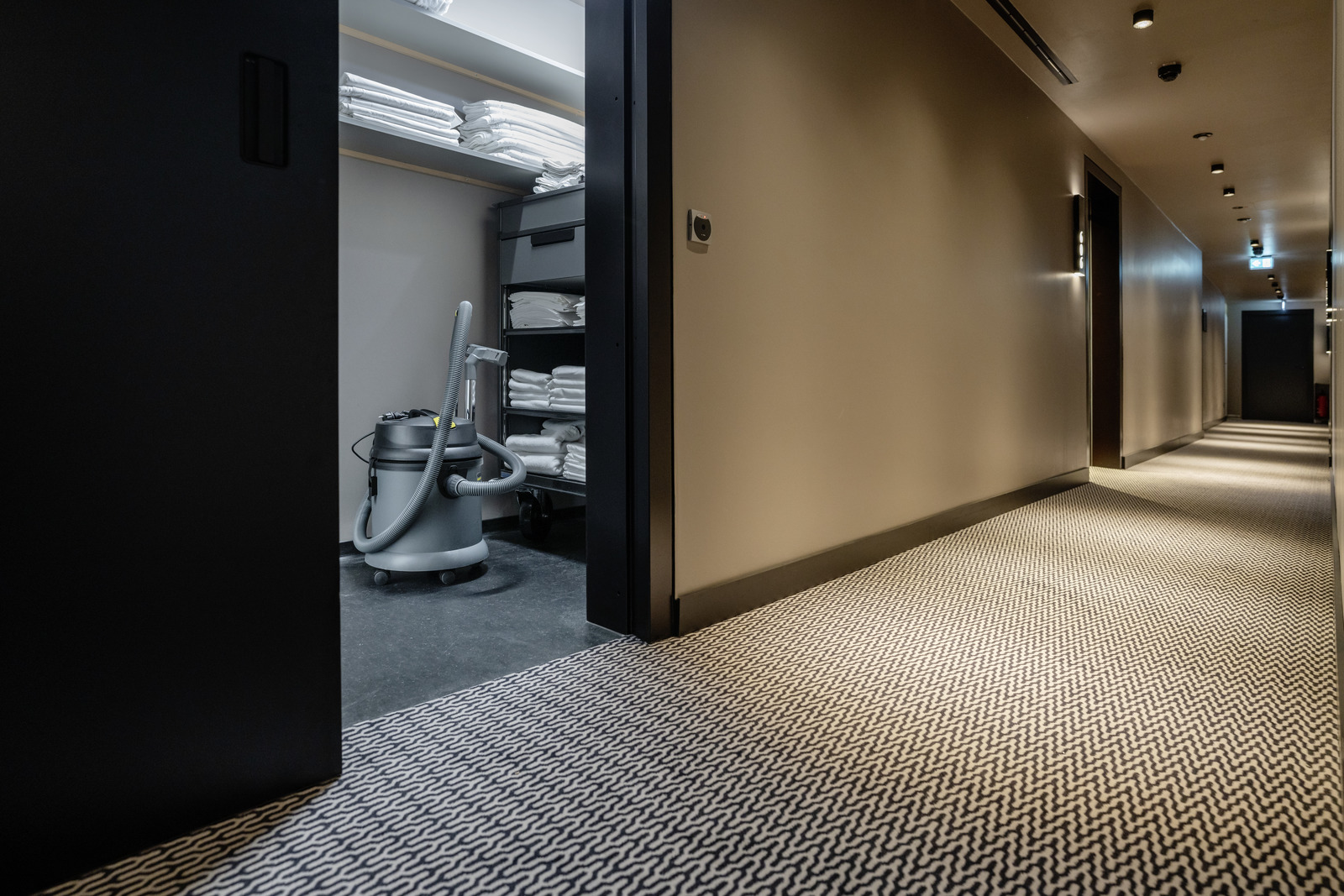 Hallway with patterned carpet leading to a storage room with shelves of folded towels and a Kärcher vacuum cleaner.