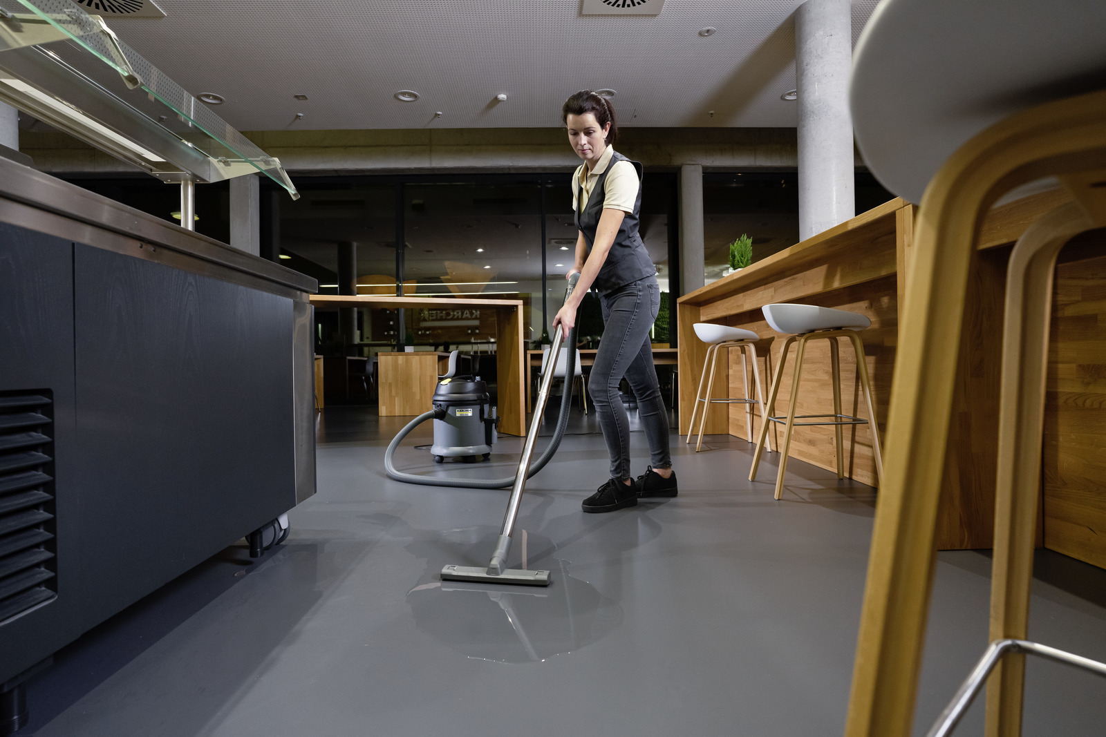 Person using a Kärcher wet and dry vacuum cleaner on a grey floor in a modern indoor setting with wooden furniture and bar stools.