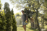 Man using Kärcher garden sprayer in sunny garden, surrounded by trees and shrubs, with a wooden shed in the background.