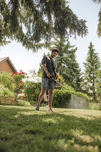 Person using Kärcher high-pressure cleaner on lawn in a garden with trees, red flowers, and a house in the background.
