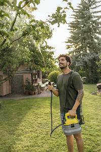 Man using Kärcher sprayer to water garden plants, standing on grass with trees and a house in the background.