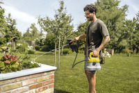 Man using Kärcher sprayer in a garden, watering plants in a raised brick planter surrounded by greenery.