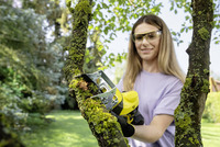 Person using a Kärcher chainsaw to cut a moss-covered tree branch in a garden setting.