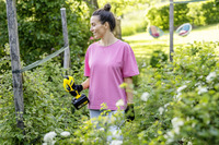 Person in pink shirt holding Kärcher battery-powered pruner in a lush garden setting, surrounded by greenery and wooden posts.