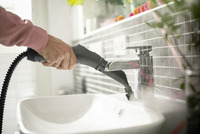 Hand using Kärcher steam cleaner on bathroom tap, with steam visible. Black tiled wall and white sink in the background.