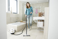 Woman cleaning bathroom floor with Kärcher steam cleaner, surrounded by modern fixtures and neutral tiles.