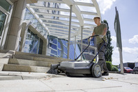 Person using a Kärcher push sweeper on outdoor stone steps near a modern glass building under a clear sky.