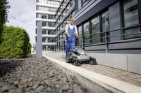 Person using a Kärcher floor sweeper on a paved path outside a modern office building, surrounded by greenery and gravel.