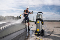 Worker cleaning a rooftop with a Kärcher high-pressure cleaner, wearing safety gear and harness under a partly cloudy sky.