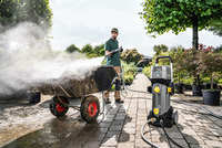 Person using Kärcher high-pressure cleaner on a wheelbarrow outdoors, surrounded by plants and trees on a paved path.
