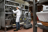 Worker cleaning industrial machine with Kärcher steam cleaner in a factory setting, wearing protective clothing and gloves.