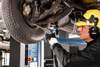Worker uses Kärcher high-pressure cleaner on car's undercarriage in a garage, wearing protective gear including helmet and earmuffs.