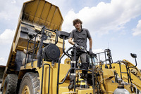 Man standing on a large yellow construction vehicle, holding a tool, with a raised dump bed in an outdoor setting under a cloudy sky.