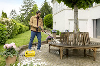 Man cleaning wooden bench with Kärcher high-pressure cleaner in a garden setting, surrounded by potted flowers and greenery.
