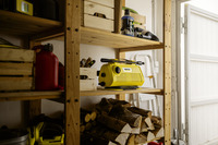 Yellow Kärcher high-pressure cleaner on wooden shelves in a garage, surrounded by logs, tools, and cleaning supplies.