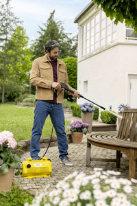 Man using Kärcher high-pressure cleaner on wooden garden furniture, surrounded by potted flowers and greenery, near a white house.