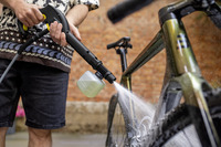 Person cleaning a bicycle with a Kärcher high-pressure cleaner, outdoors against a brick wall background.
