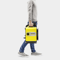 Person in grey jumper and jeans carrying a yellow Kärcher high-pressure cleaner, with grey trainers, against a plain background.